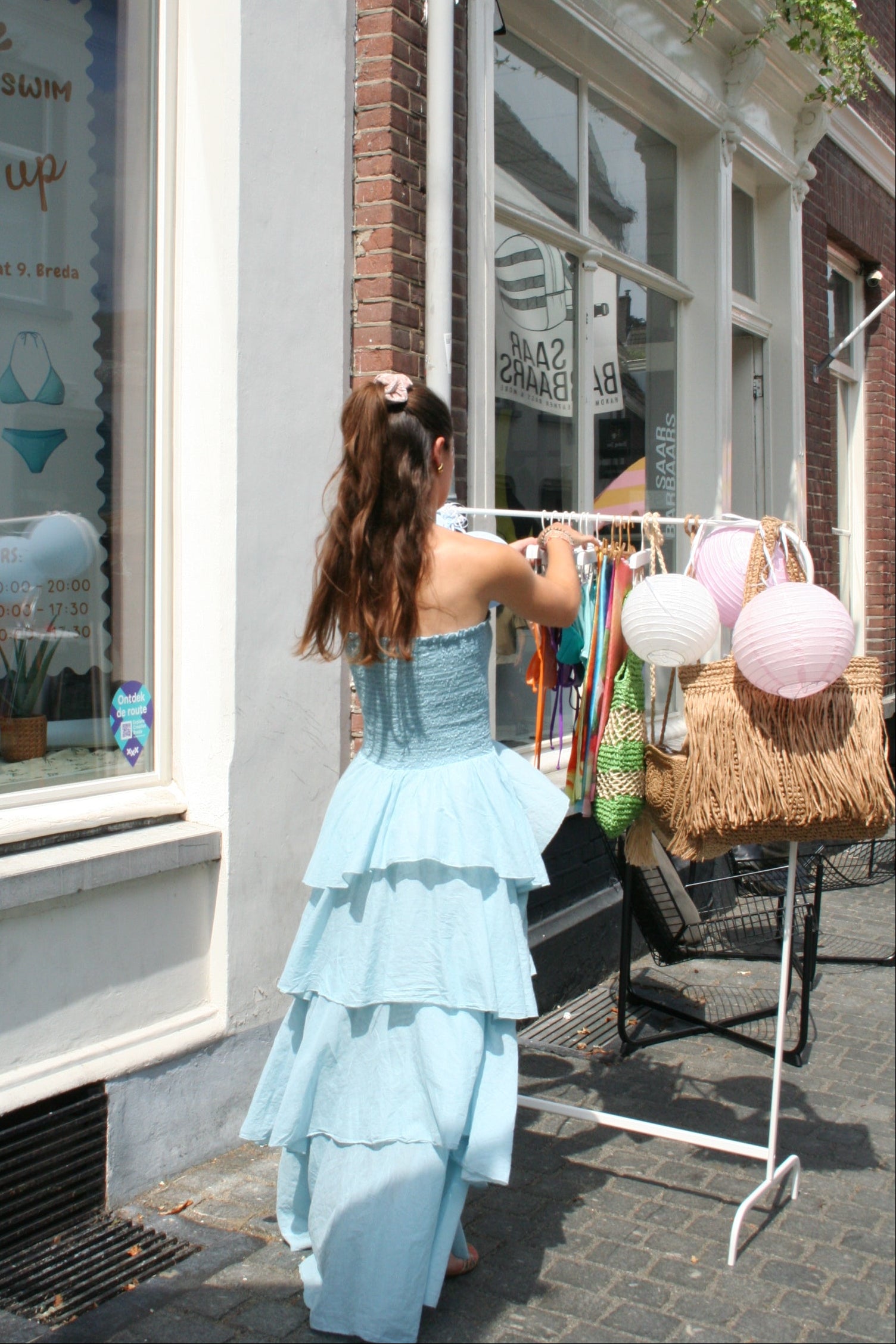 A woman wearing a light blue tiered ruffled maxi dress stands on a street corner next to a shop window, holding a white plastic bag. She is positioned next to a straw basket and pink and white paper lanterns.